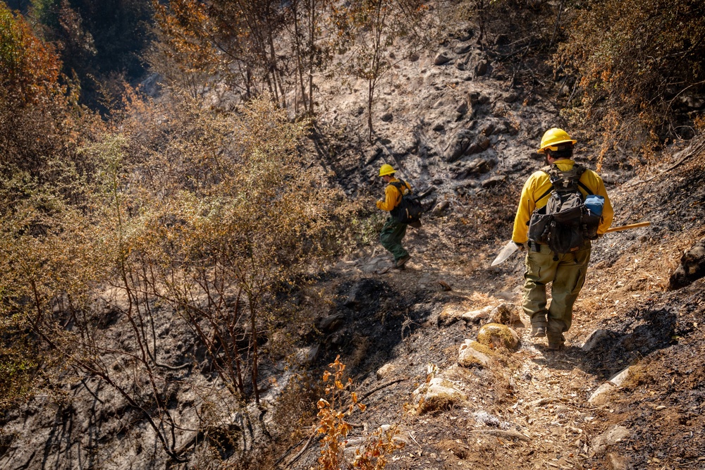 Two wildland firefighters in standard yellow Nomex gear and hard hats traverse a steep, fire-scarred hillside. The surrounding terrain shows extensive burn damage with charred soil, scorched vegetation, and blackened tree trunks. The firefighters appear to be conducting post-fire mop-up or patrol operations along a ridge or drainage.