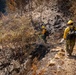 Two wildland firefighters in standard yellow Nomex gear and hard hats traverse a steep, fire-scarred hillside. The surrounding terrain shows extensive burn damage with charred soil, scorched vegetation, and blackened tree trunks. The firefighters appear to be conducting post-fire mop-up or patrol operations along a ridge or drainage.