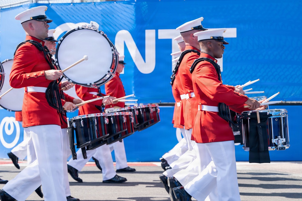 A military marching band, identifiable as U.S. Marines based on their distinctive red tunics and white covers (caps), performs in a parade or public event. The musicians are playing various drums including a bass drum and snare/tenor drums while marching in formation. A blue banner with partial white lettering is visible in the background.