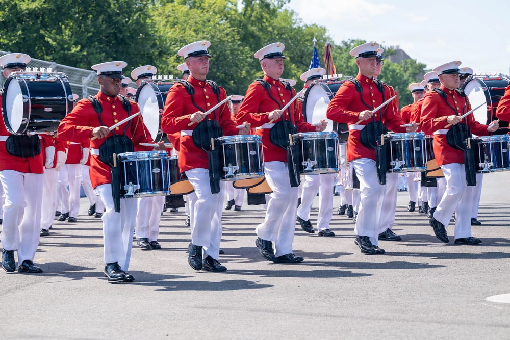 A military marching band is performing in formation on an outdoor paved surface. The musicians are dressed in distinctive red tunics, white trousers, and white peaked caps, consistent with U.S. Marine Corps ceremonial dress uniforms. They are playing snare drums and bass drums while marching in synchronized formation, with an American flag visible in the background.