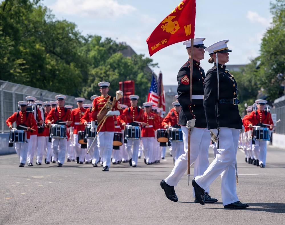 A United States Marine Corps marching band and color guard are performing in a parade, likely in Washington D.C. Two Marines in dark dress blues lead the procession, with one carrying a red USMC guidon flag marked 'WASH DC'. Behind them, a large formation of band members in red and white dress uniforms march while playing snare drums and other instruments.