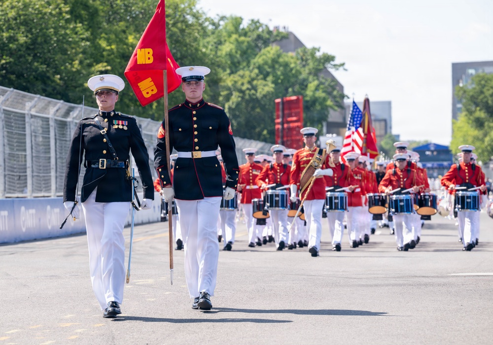 A U.S. Marine Corps marching band parade is taking place on a paved street or circuit. Two lead Marines in dress blues march at the front, one carrying a red 'MB' (Marine Band) guidon flag, while a large formation of band members in red uniforms with drums follows behind. An American flag and additional Marine Corps colors are visible in the procession.