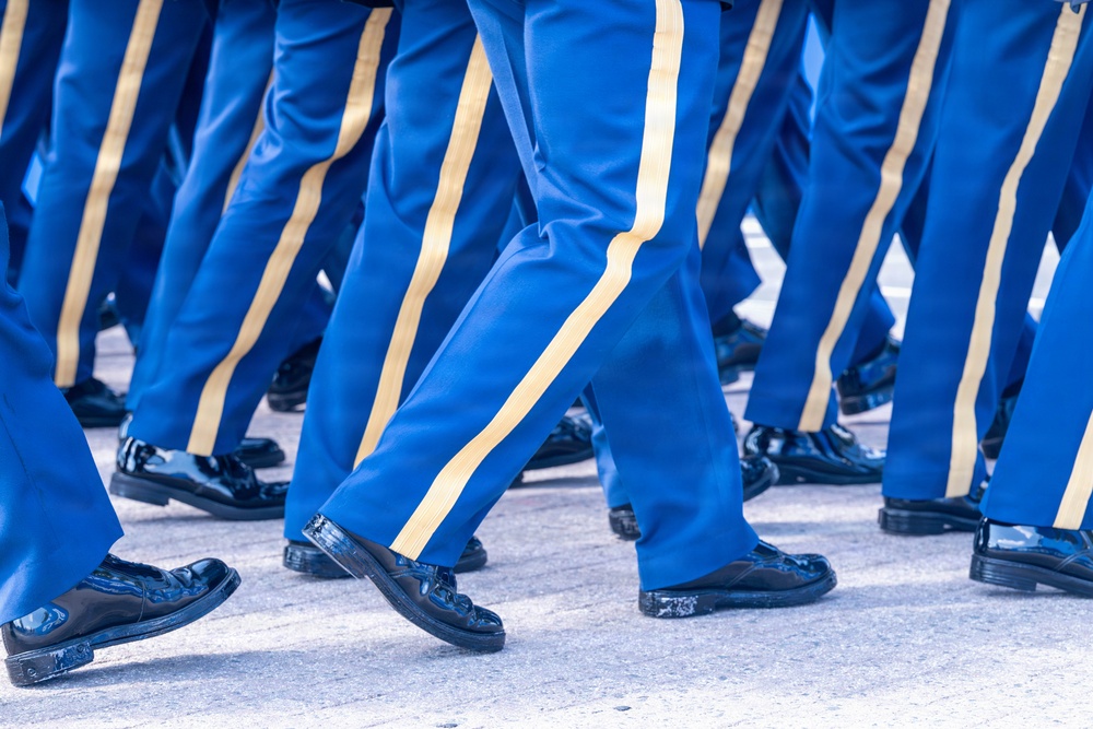 The image shows a close-up view of multiple individuals marching in formation, wearing matching blue dress uniforms with gold/yellow side stripes on the trousers. Only the lower half of the participants is visible, from approximately the waist down. They are wearing highly polished black dress shoes, consistent with a formal parade or ceremonial march.