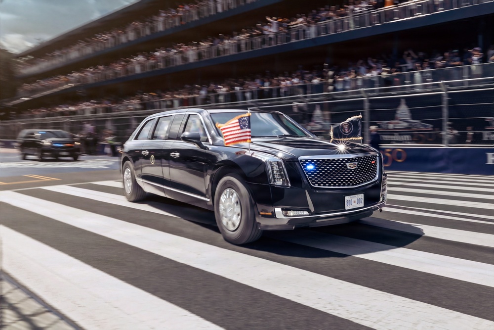 A large black armored presidential limousine, commonly known as 'The Beast,' is traveling along a parade route flanked by bleachers filled with spectators. The vehicle displays an American flag and what appears to be a presidential seal flag, along with blue emergency lights active on the grille. A follow-up vehicle is visible behind it, consistent with a standard presidential motorcade formation.