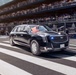 A large black armored presidential limousine, commonly known as 'The Beast,' is traveling along a parade route flanked by bleachers filled with spectators. The vehicle displays an American flag and what appears to be a presidential seal flag, along with blue emergency lights active on the grille. A follow-up vehicle is visible behind it, consistent with a standard presidential motorcade formation.