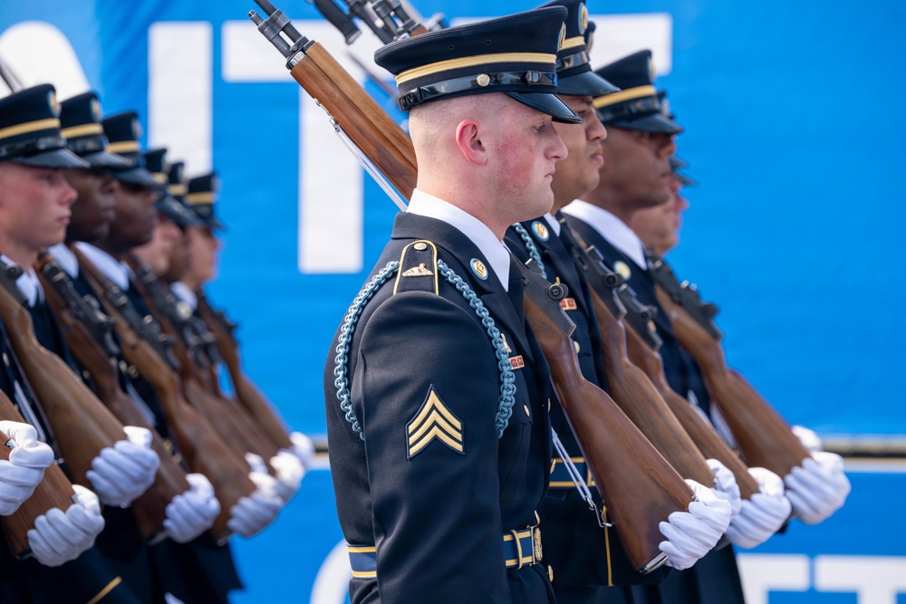 A formation of U.S. Army soldiers in dress uniform are standing at attention during a formal military ceremony or parade. The soldiers are equipped with rifles carried on their shoulders and are wearing white gloves, ceremonial dress blues, and service caps. The foreground soldier displays Sergeant rank chevrons and decorative aiguillette, consistent with a ceremonial honor guard unit.
