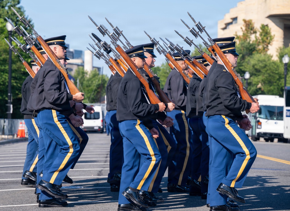 A column of military honor guard soldiers marches in precise formation along a paved street. They are dressed in formal ceremonial uniforms featuring blue trousers with gold stripes, dark jackets, and black peaked caps. Each soldier carries a rifle with a fixed bayonet, held at a uniform angle, indicating a formal parade or ceremonial event.