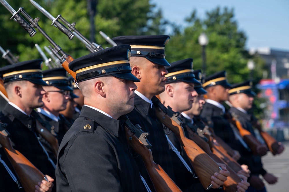 A formation of military personnel in formal dress uniforms are standing at attention during what appears to be a ceremonial parade or review. The soldiers are carrying rifles with bayonets fixed, suggesting a honor guard or ceremonial unit. The setting appears to be an outdoor public venue with trees and urban infrastructure visible in the background.