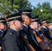 A formation of military personnel in formal dress uniforms are standing at attention during what appears to be a ceremonial parade or review. The soldiers are carrying rifles with bayonets fixed, suggesting a honor guard or ceremonial unit. The setting appears to be an outdoor public venue with trees and urban infrastructure visible in the background.