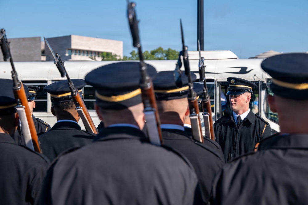 A military or ceremonial honor guard unit is assembled in formation, holding rifles with fixed bayonets at shoulder arms. A single soldier facing the group appears to be an officer or non-commissioned officer conducting an inspection or giving instructions. The setting appears to be an outdoor area near a bus and institutional buildings under clear blue skies.