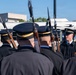 A military or ceremonial honor guard unit is assembled in formation, holding rifles with fixed bayonets at shoulder arms. A single soldier facing the group appears to be an officer or non-commissioned officer conducting an inspection or giving instructions. The setting appears to be an outdoor area near a bus and institutional buildings under clear blue skies.
