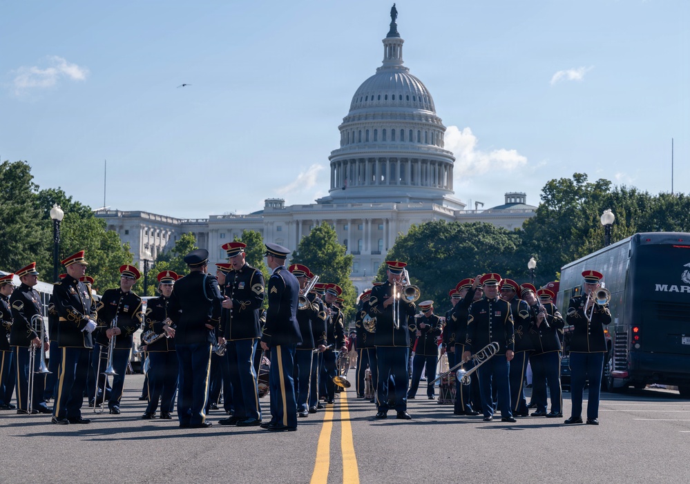 A military marching band is assembled on a street directly in front of the United States Capitol Building in Washington, D.C. The musicians are wearing dark dress uniforms with distinctive red-plumed caps and are holding various brass instruments including trombones, tubas, and trumpets. The scene appears to be a pre-parade or ceremonial gathering on a clear, sunny day.