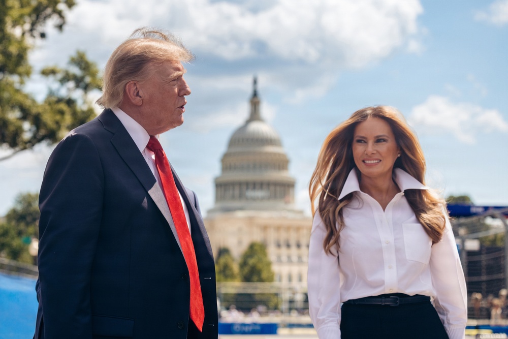 Two individuals are standing outdoors in front of the United States Capitol building in Washington, D.C. The man on the left is wearing a dark navy suit with a red tie, while the woman on the right is wearing a white button-down shirt and dark skirt. The setting appears to be a daytime public event with clear skies and partial cloud cover.