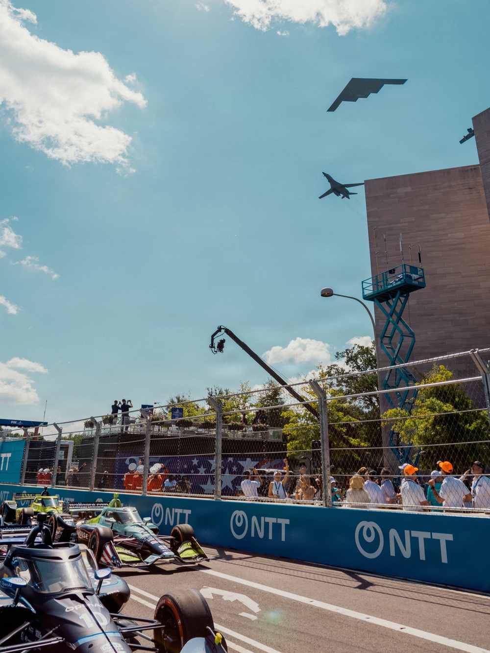 An IndyCar racing event is taking place on a street circuit, with multiple open-wheel race cars lined up on pit lane or the starting grid. A military flyover is occurring simultaneously, with at least three aircraft visible in the sky including what appears to be a B-2 Spirit stealth bomber, a B-1 Lancer, and a third aircraft. Spectators and crew members line the pit wall and grandstand areas, watching both the aircraft and the race proceedings.