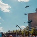 An IndyCar racing event is taking place on a street circuit, with multiple open-wheel race cars lined up on pit lane or the starting grid. A military flyover is occurring simultaneously, with at least three aircraft visible in the sky including what appears to be a B-2 Spirit stealth bomber, a B-1 Lancer, and a third aircraft. Spectators and crew members line the pit wall and grandstand areas, watching both the aircraft and the race proceedings.