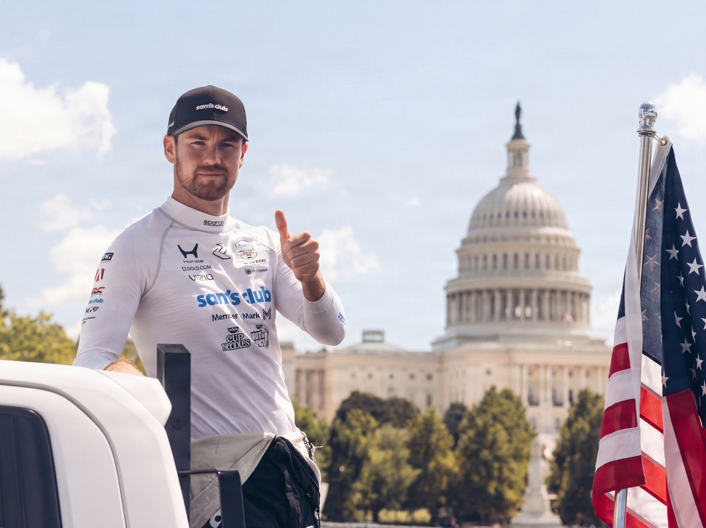 A racing driver wearing a white Sam's Club-sponsored racing suit and matching cap gives a thumbs-up gesture in front of the United States Capitol building in Washington, D.C. The driver's uniform displays multiple sponsor logos including Honda, Sam's Club, Member Mark, Cup Noodles, and others. An American flag is visible on the right side of the frame.