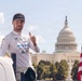 A racing driver wearing a white Sam's Club-sponsored racing suit and matching cap gives a thumbs-up gesture in front of the United States Capitol building in Washington, D.C. The driver's uniform displays multiple sponsor logos including Honda, Sam's Club, Member Mark, Cup Noodles, and others. An American flag is visible on the right side of the frame.