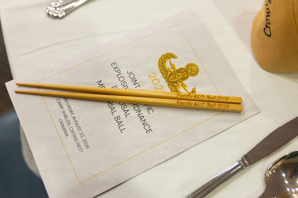 The image shows a formal place setting at a military ball event, featuring engraved wooden chopsticks resting on a printed program. The program and chopsticks both reference the 'Pacific EOD Ball 2026,' held on Saturday, August 22, 2026, at Camp Shields, Crow's Nest, Okinawa. The event is identified as a Joint Pacific Explosive Ordnance Disposal Memorial Ball.