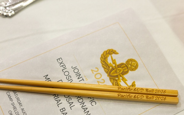 The image shows a formal place setting at a military ball event, featuring engraved wooden chopsticks resting on a printed program. The program and chopsticks both reference the 'Pacific EOD Ball 2026,' held on Saturday, August 22, 2026, at Camp Shields, Crow's Nest, Okinawa. The event is identified as a Joint Pacific Explosive Ordnance Disposal Memorial Ball.