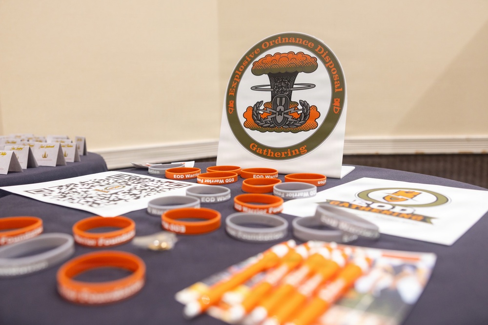 A display table set up for an Explosive Ordnance Disposal (EOD) Gathering event, featuring branded merchandise and informational materials. The table is covered with orange and gray silicone wristbands labeled 'EOD Warrior Foundation,' along with lanyards, QR code sheets, and a prominent circular EOD Gathering logo sign. The setup appears to be a registration or merchandise booth at a military or veteran-related event.