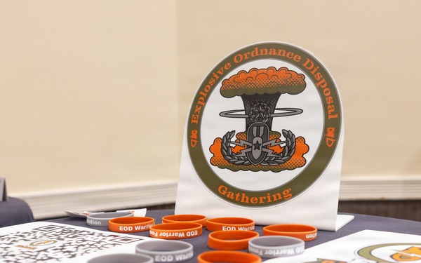 A display table set up for an Explosive Ordnance Disposal (EOD) Gathering event, featuring branded merchandise and informational materials. The table is covered with orange and gray silicone wristbands labeled 'EOD Warrior Foundation,' along with lanyards, QR code sheets, and a prominent circular EOD Gathering logo sign. The setup appears to be a registration or merchandise booth at a military or veteran-related event.