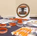 A display table set up for an Explosive Ordnance Disposal (EOD) Gathering event, featuring branded merchandise and informational materials. The table is covered with orange and gray silicone wristbands labeled 'EOD Warrior Foundation,' along with lanyards, QR code sheets, and a prominent circular EOD Gathering logo sign. The setup appears to be a registration or merchandise booth at a military or veteran-related event.
