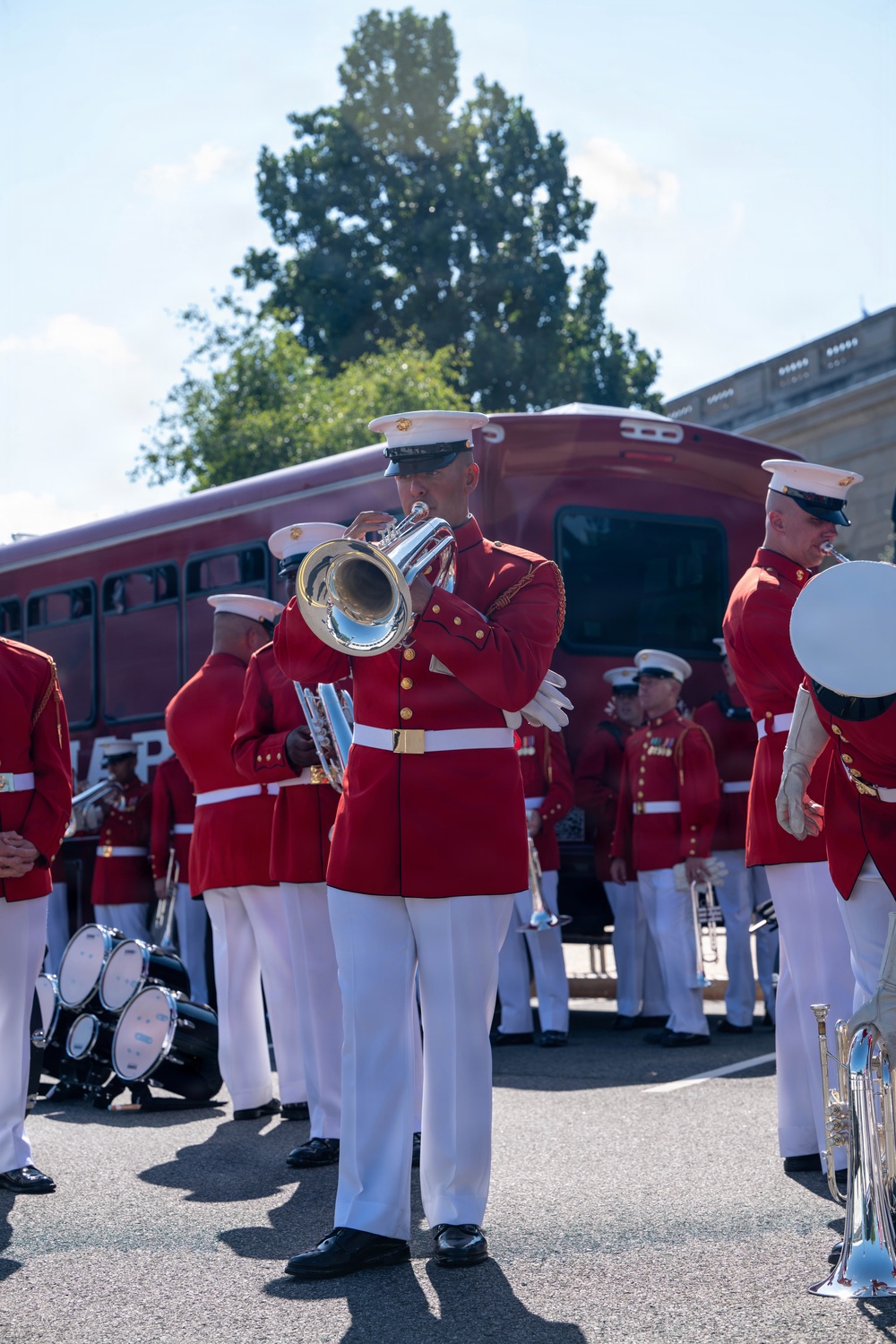 A U.S. Marine Corps marching band is assembled outdoors, performing in their distinctive scarlet and white dress uniforms. The musicians are playing brass instruments including a mellophone/marching French horn and what appears to be a euphonium/baritone horn. The setting appears to be a paved area near a red passenger rail car and a stone building, suggesting a formal ceremonial or public event.
