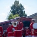 A U.S. Marine Corps marching band is assembled outdoors, performing in their distinctive scarlet and white dress uniforms. The musicians are playing brass instruments including a mellophone/marching French horn and what appears to be a euphonium/baritone horn. The setting appears to be a paved area near a red passenger rail car and a stone building, suggesting a formal ceremonial or public event.
