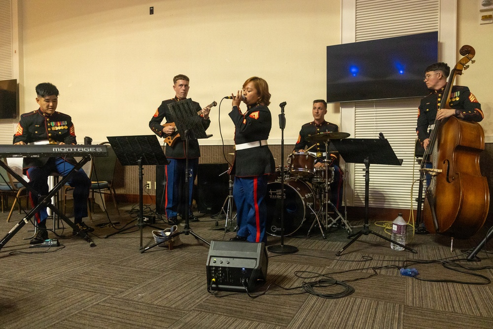 A U.S. Marine Corps musical ensemble performs in what appears to be an indoor venue or event hall. Five musicians in dress blue uniforms are playing various instruments while one female Marine serves as the lead vocalist. The group includes keyboard, guitar, drums, double bass, and a singer, suggesting a jazz or contemporary small ensemble format.