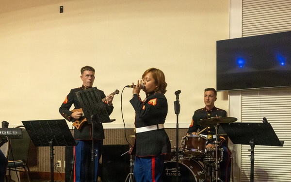 A U.S. Marine Corps musical ensemble performs in what appears to be an indoor venue or event hall. Five musicians in dress blue uniforms are playing various instruments while one female Marine serves as the lead vocalist. The group includes keyboard, guitar, drums, double bass, and a singer, suggesting a jazz or contemporary small ensemble format.