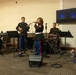 A U.S. Marine Corps musical ensemble performs in what appears to be an indoor venue or event hall. Five musicians in dress blue uniforms are playing various instruments while one female Marine serves as the lead vocalist. The group includes keyboard, guitar, drums, double bass, and a singer, suggesting a jazz or contemporary small ensemble format.
