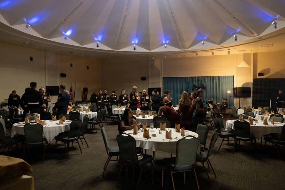 A formal military banquet or ball is taking place in a large circular ballroom with a distinctive domed ceiling illuminated by blue uplighting. Multiple military personnel in dress uniforms, likely U.S. Marines based on the distinctive dark dress blues, are mingling with civilian guests in formal attire. The room is set with round banquet tables covered in white linens with folded napkins, suggesting a formal dinner event that is either just beginning or in a pre-dinner reception phase.