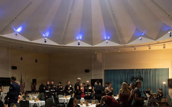 A formal military banquet or ball is taking place in a large circular ballroom with a distinctive domed ceiling illuminated by blue uplighting. Multiple military personnel in dress uniforms, likely U.S. Marines based on the distinctive dark dress blues, are mingling with civilian guests in formal attire. The room is set with round banquet tables covered in white linens with folded napkins, suggesting a formal dinner event that is either just beginning or in a pre-dinner reception phase.