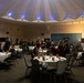 A formal military banquet or ball is taking place in a large circular ballroom with a distinctive domed ceiling illuminated by blue uplighting. Multiple military personnel in dress uniforms, likely U.S. Marines based on the distinctive dark dress blues, are mingling with civilian guests in formal attire. The room is set with round banquet tables covered in white linens with folded napkins, suggesting a formal dinner event that is either just beginning or in a pre-dinner reception phase.