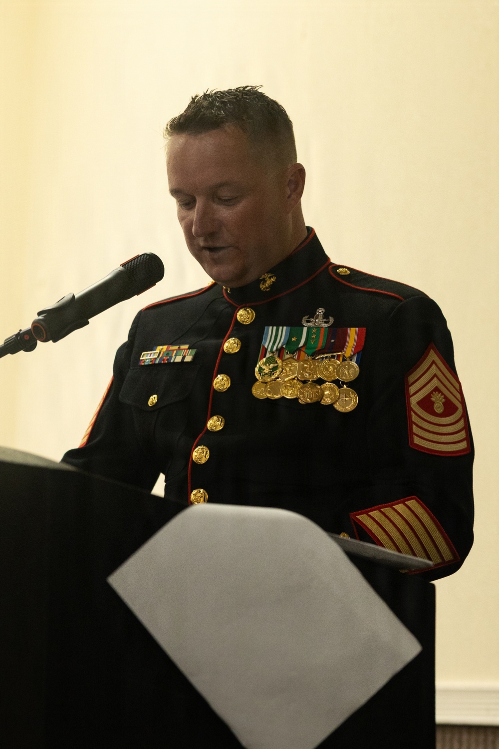 A U.S. Marine Corps senior enlisted leader in dress blues uniform is speaking at a podium during a formal ceremony or event. The individual displays an extensive array of medals, ribbons, and rank insignia consistent with a highly decorated senior non-commissioned officer. They are reading from papers placed on the lectern while speaking into a microphone.