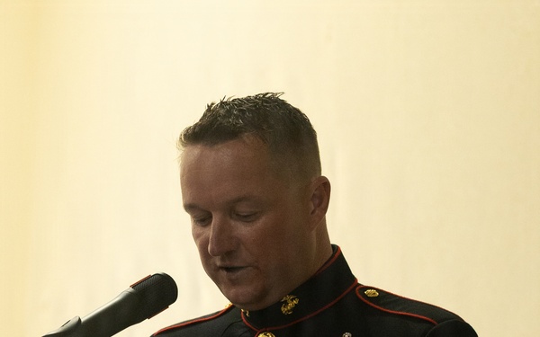 A U.S. Marine Corps senior enlisted leader in dress blues uniform is speaking at a podium during a formal ceremony or event. The individual displays an extensive array of medals, ribbons, and rank insignia consistent with a highly decorated senior non-commissioned officer. They are reading from papers placed on the lectern while speaking into a microphone.