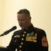 A U.S. Marine Corps senior enlisted leader in dress blues uniform is speaking at a podium during a formal ceremony or event. The individual displays an extensive array of medals, ribbons, and rank insignia consistent with a highly decorated senior non-commissioned officer. They are reading from papers placed on the lectern while speaking into a microphone.