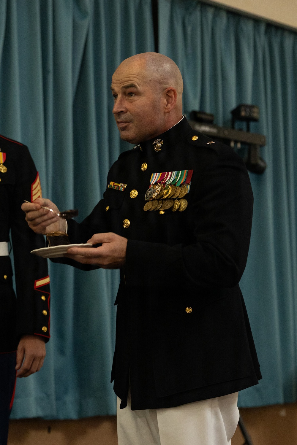 A bald male officer in United States Marine Corps dress blues uniform stands holding a small plate and an object during what appears to be a formal military ceremony or event. He wears a single star on his shoulder indicating the rank of Brigadier General, along with multiple rows of medals and ribbons on his chest. The setting appears to be an indoor venue with a blue curtain backdrop.