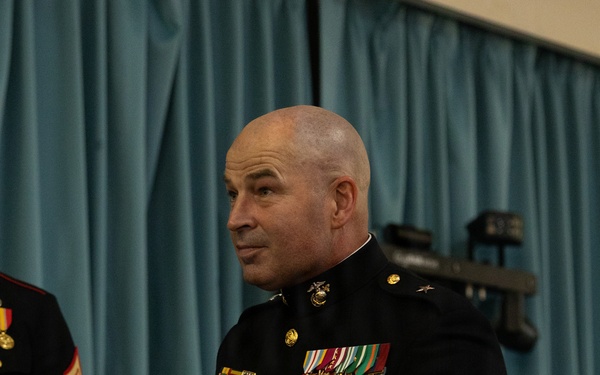 A bald male officer in United States Marine Corps dress blues uniform stands holding a small plate and an object during what appears to be a formal military ceremony or event. He wears a single star on his shoulder indicating the rank of Brigadier General, along with multiple rows of medals and ribbons on his chest. The setting appears to be an indoor venue with a blue curtain backdrop.