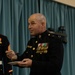 A bald male officer in United States Marine Corps dress blues uniform stands holding a small plate and an object during what appears to be a formal military ceremony or event. He wears a single star on his shoulder indicating the rank of Brigadier General, along with multiple rows of medals and ribbons on his chest. The setting appears to be an indoor venue with a blue curtain backdrop.