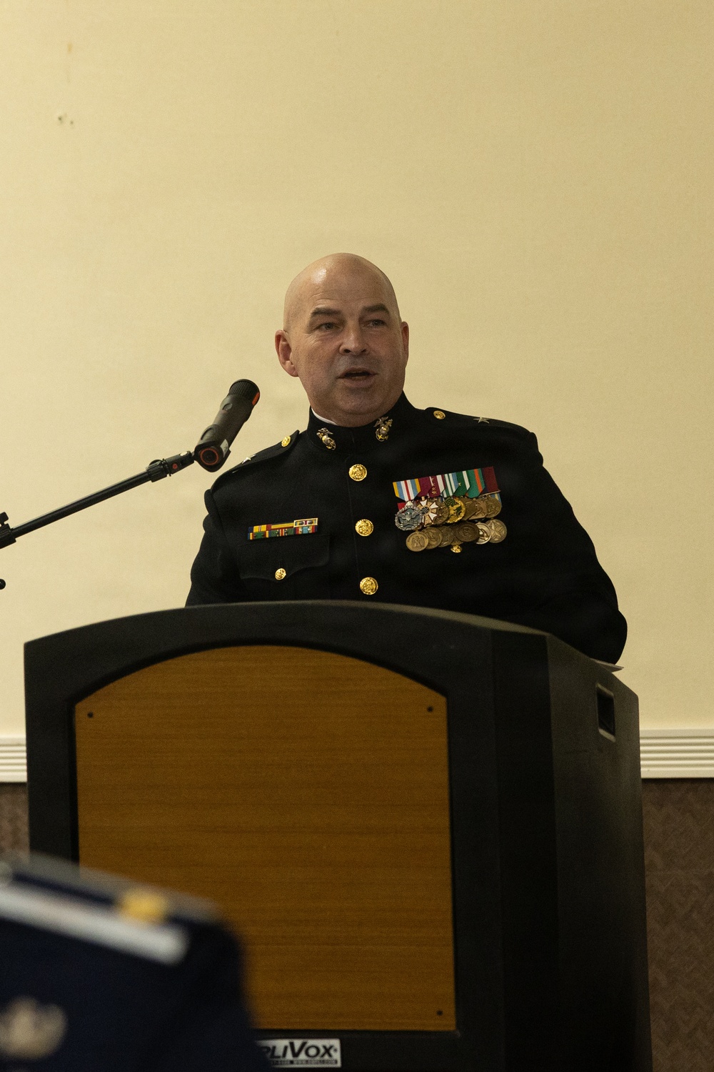 A bald male military officer in U.S. Marine Corps dress blues uniform is speaking at a wooden podium equipped with a microphone. He is wearing multiple medals, ribbons, and gold buttons characteristic of formal Marine Corps dress uniform. The setting appears to be an indoor formal ceremony or official event.