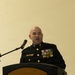 A bald male military officer in U.S. Marine Corps dress blues uniform is speaking at a wooden podium equipped with a microphone. He is wearing multiple medals, ribbons, and gold buttons characteristic of formal Marine Corps dress uniform. The setting appears to be an indoor formal ceremony or official event.