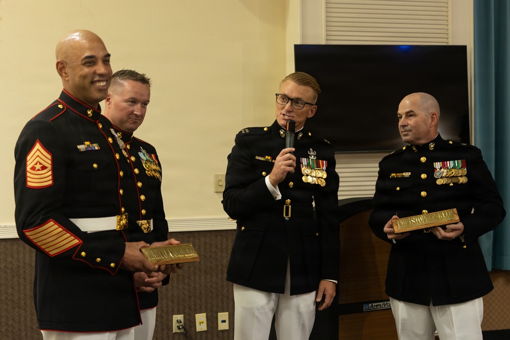 Four U.S. Marine Corps personnel in dress blue uniforms are gathered at what appears to be a formal military ceremony or retirement event. Two of the individuals are holding engraved name plaques, while a third speaks into a microphone. The setting appears to be an indoor venue with a television screen visible in the background.