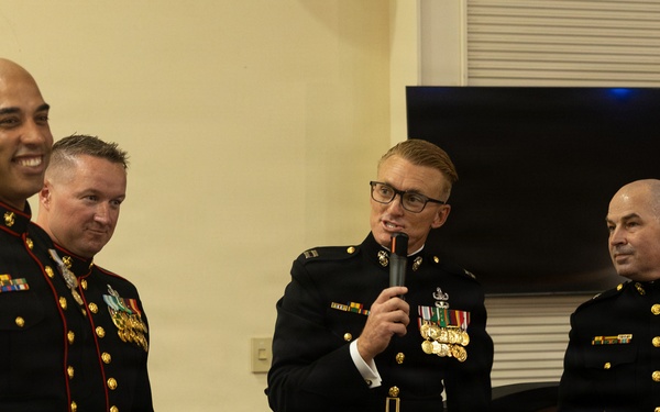 Four U.S. Marine Corps personnel in dress blue uniforms are gathered at what appears to be a formal military ceremony or retirement event. Two of the individuals are holding engraved name plaques, while a third speaks into a microphone. The setting appears to be an indoor venue with a television screen visible in the background.