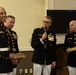 Four U.S. Marine Corps personnel in dress blue uniforms are gathered at what appears to be a formal military ceremony or retirement event. Two of the individuals are holding engraved name plaques, while a third speaks into a microphone. The setting appears to be an indoor venue with a television screen visible in the background.
