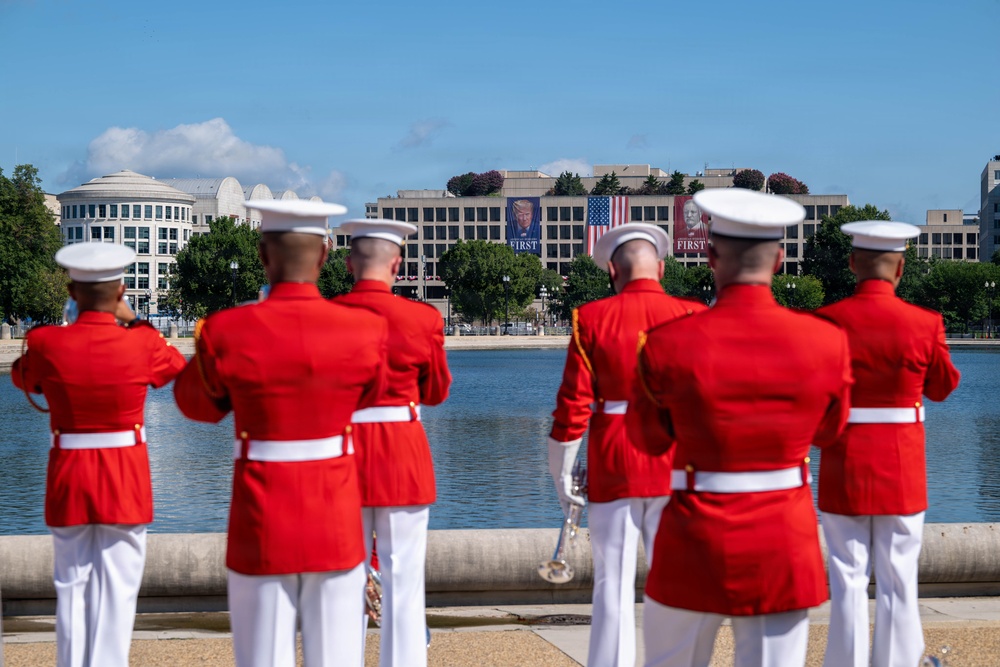 A group of U.S. Marine Corps musicians in ceremonial dress uniforms stand at attention near a reflecting pool in Washington, D.C. In the background, a large government building displays inauguration-related banners featuring portraits and American flags. The scene appears to be part of an official inaugural or ceremonial event.