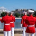 A group of U.S. Marine Corps musicians in ceremonial dress uniforms stand at attention near a reflecting pool in Washington, D.C. In the background, a large government building displays inauguration-related banners featuring portraits and American flags. The scene appears to be part of an official inaugural or ceremonial event.