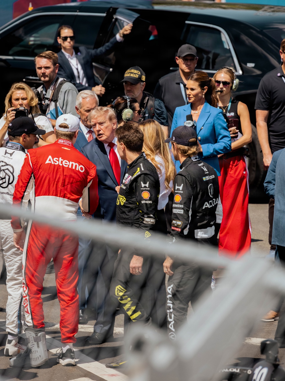 A group of people gathered on what appears to be a racing event pit lane or starting grid, including several individuals wearing racing suits with visible sponsor logos such as Astemo, Acura, Shell, Siemens, and Honda. A man in a blue suit with a red tie is prominently positioned in the center of the group, surrounded by racing personnel, media, and security. A woman in a blue blazer holding a FOX microphone appears to be conducting interviews nearby.