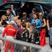 A group of people gathered on what appears to be a racing event pit lane or starting grid, including several individuals wearing racing suits with visible sponsor logos such as Astemo, Acura, Shell, Siemens, and Honda. A man in a blue suit with a red tie is prominently positioned in the center of the group, surrounded by racing personnel, media, and security. A woman in a blue blazer holding a FOX microphone appears to be conducting interviews nearby.