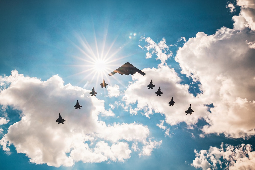 A dramatic aerial formation photograph taken from below, showing a large flying-wing stealth bomber (consistent with a B-2 Spirit) flanked by multiple smaller twin-tail fighter jets (consistent with F-22 Raptors) flying in a V-shaped escort formation. The image is backlit by a bright sunburst against a blue sky with large cumulus clouds, rendering all aircraft as dark silhouettes. The composition creates a striking contrast between the aircraft silhouettes and the luminous sky.